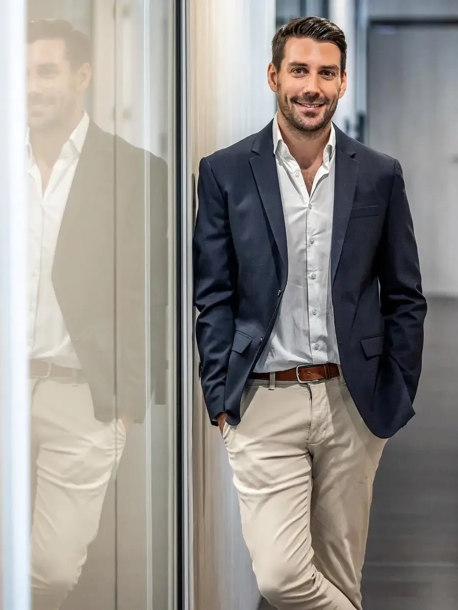 A smiling professional man in a dark jacket and light pants leans against a wall.